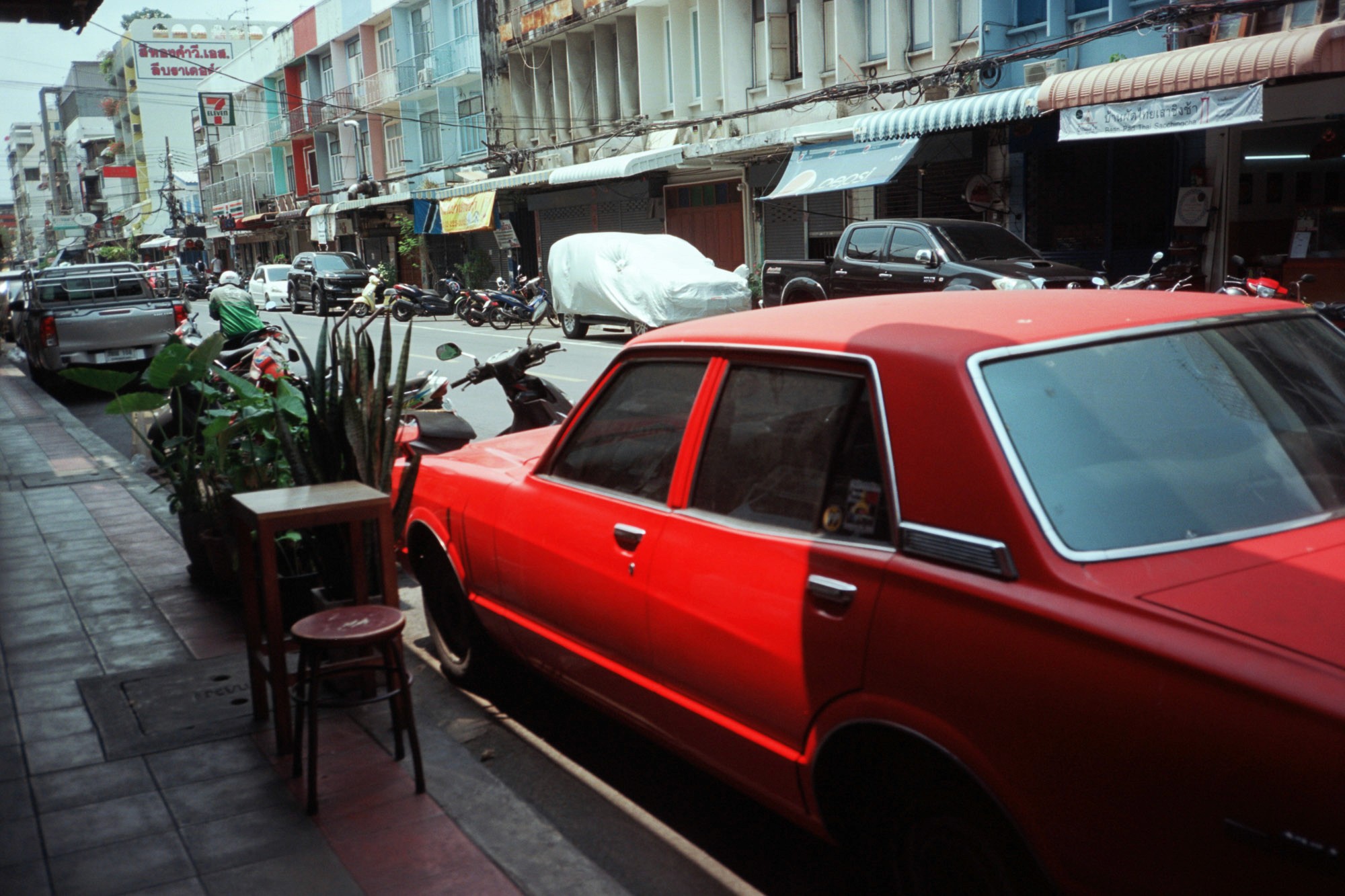 A vibrant red vintage car is parked on a busy urban street, surrounded by other vehicles and motorcycles, with pedestrians walking along tiled sidewalks lined with potted plants and small café tables.