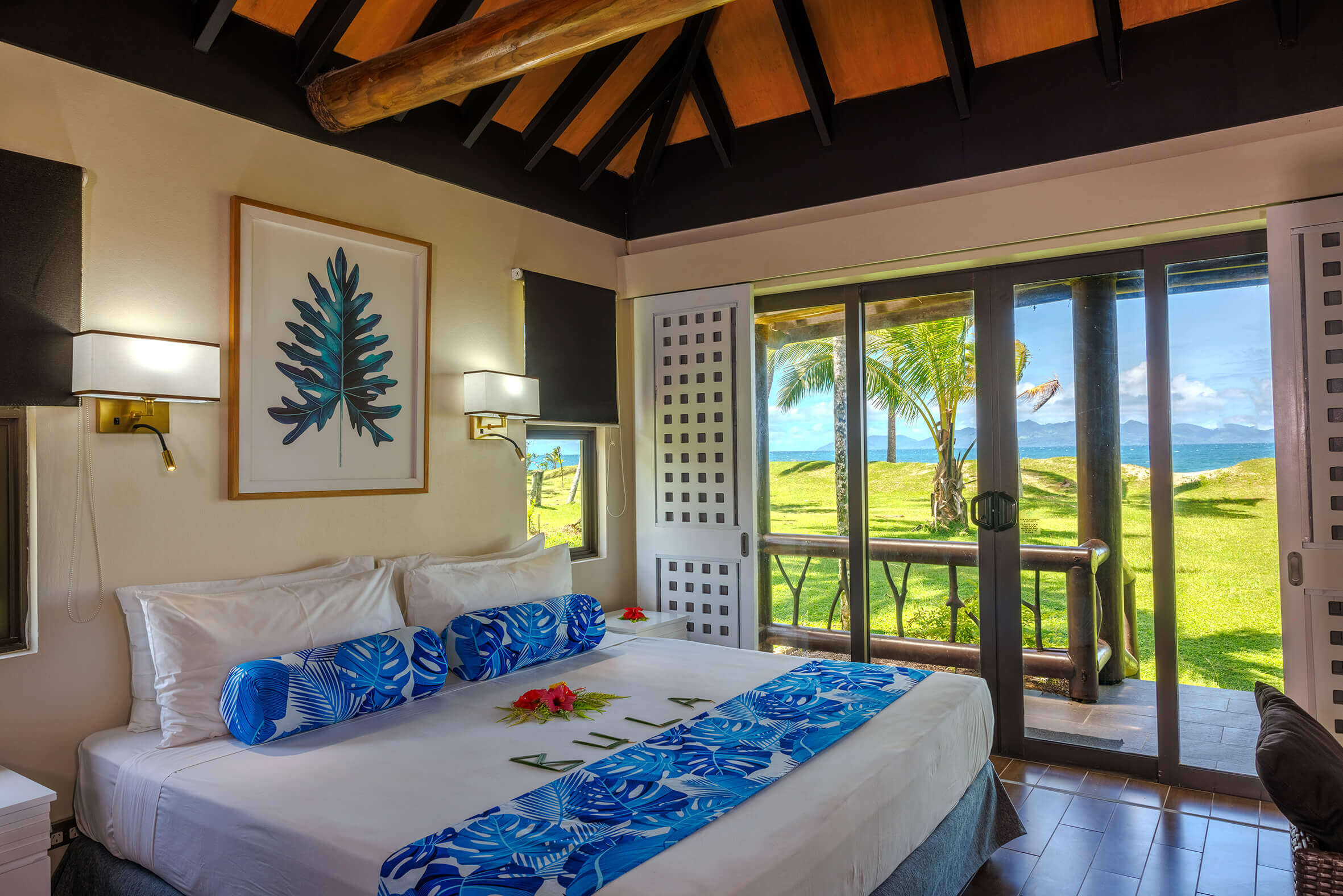 Inviting beachfront bure at Uprising Beach Resort Fiji with a decorated bed and a view of green lawn, palm trees, and the ocean.