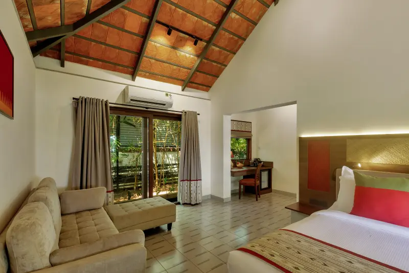 Junior Suite living area at Vedic Village Kodungallur with vaulted terracotta ceiling, beige sofa, garden-view sliding doors and red-accent bed