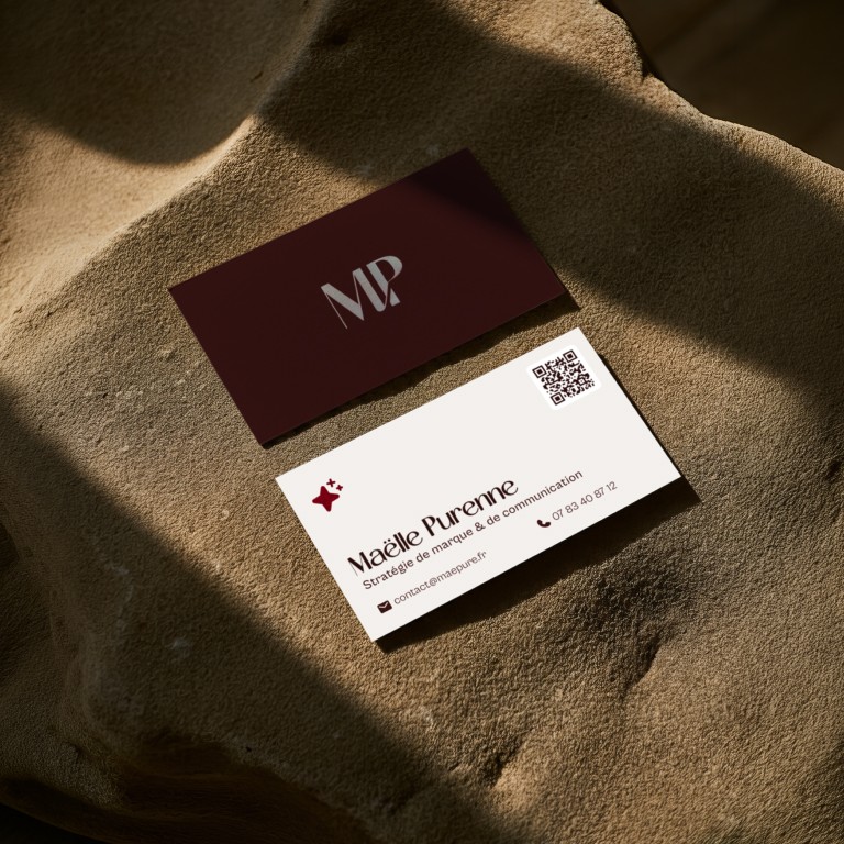 Mockup en situation réelle des cartes de visite de Maëlle Purenne, créées par Diana SOM. Posées sur une roche beige texturée sous un jeu d'ombres et de lumières naturelles, le visuel montre le recto et le verso de la carte : l'un au fond bordeaux minimaliste avec le logo MP beige métallisé, l'autre sur fond crème affichant le nom, les expertises “Stratégie de marque & de communication”, les coordonnées et un QR code renvoyant vers son site internet.