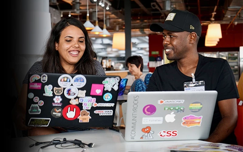 Two people seated at a table in a casual, modern indoor workspace, each working on an open laptop covered in colourful tech‑related stickers and logos. One laptop is black and heavily decorated with programming and developer community stickers, while the other is silver with stickers related to coding, JavaScript, and CSS. The person on the right is wearing a black T‑shirt and a black baseball cap, and the person on the left is wearing a dark striped top. Overhead pendant lights, exposed ceiling elements, and other people working in the background suggest a collaborative coworking space or tech meetup environment.