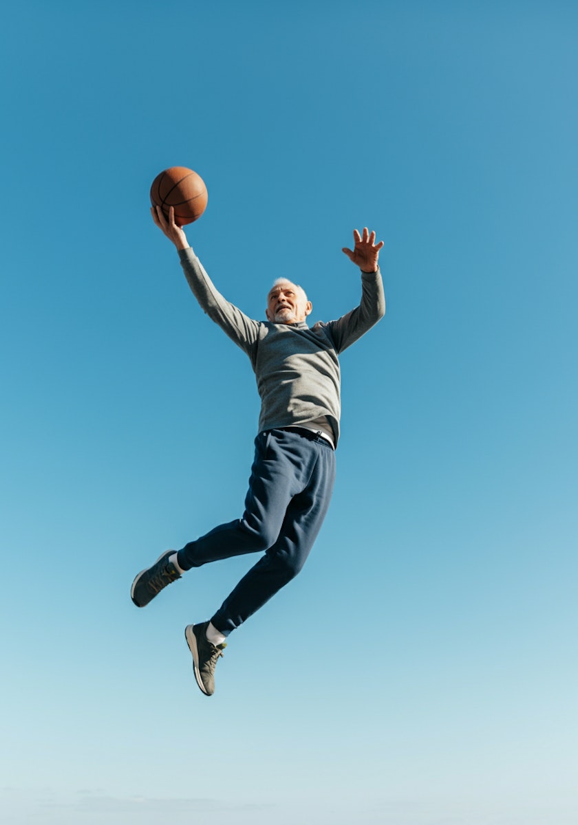 Elderly man jumping mid-air with a basketball, showcasing energy and athleticism against a clear blue sky.