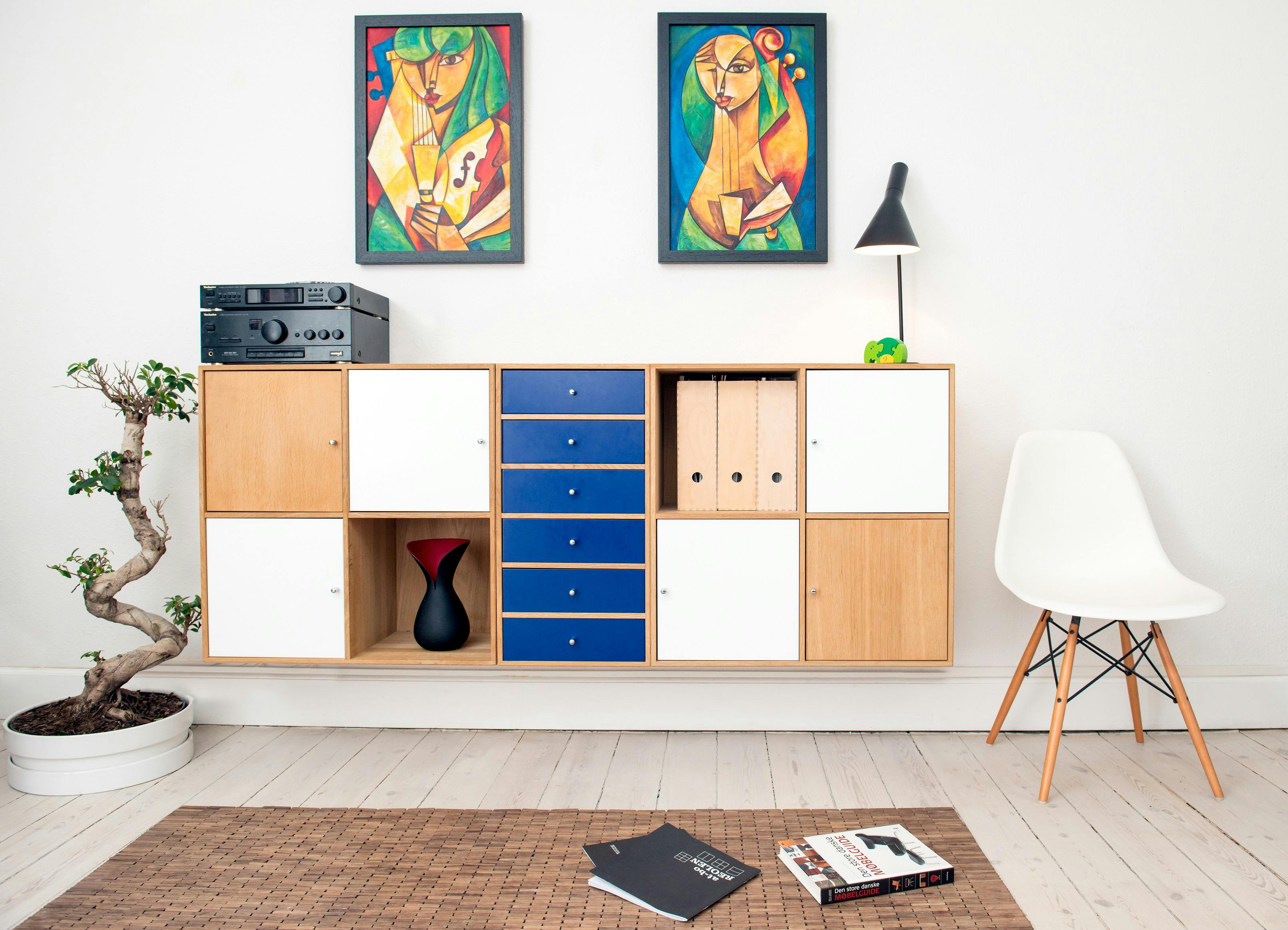Modern living room with a wooden and blue cabinet, colorful abstract paintings above. A white chair and bonsai add elegance, books scattered on the floor.