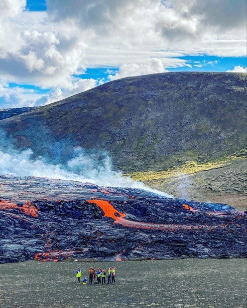 Scientists stand small in the foreground of a massive lava flow in Reykjanes, Iceland, preparing to study the newly erupting volcano. Green, mossy hills tower in the background against a blue sky, as lava tongues begin to form into the Icelandic valley.