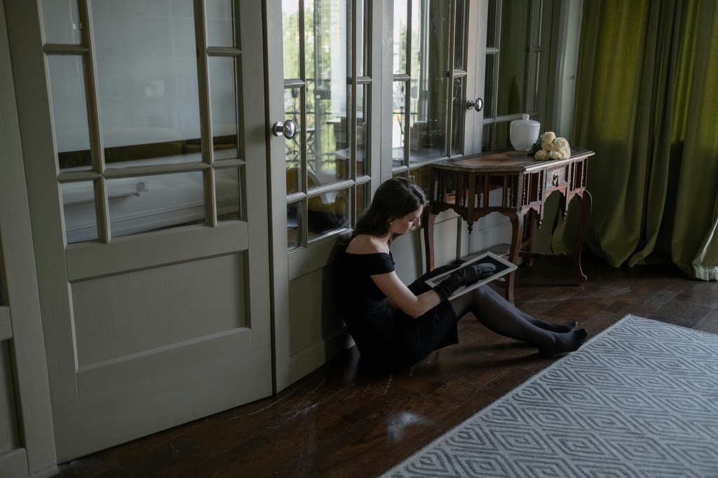 A woman in a black dress sits on the floor by French doors, intently reading a book, creating a serene and introspective atmosphere in a softly lit room with classic decor.