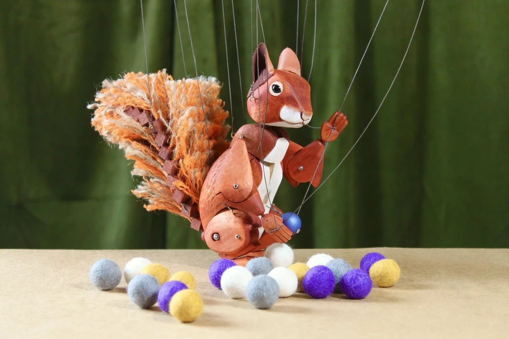 A marionette of a Squirrel stands in front of a green velvet curtain surrounded by multicoloured felt balls