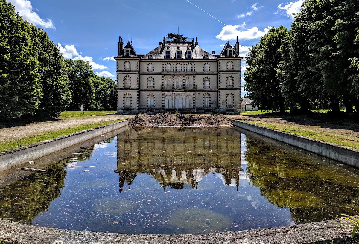A French château reflected in a long rectangular pond between rows of tall trees.