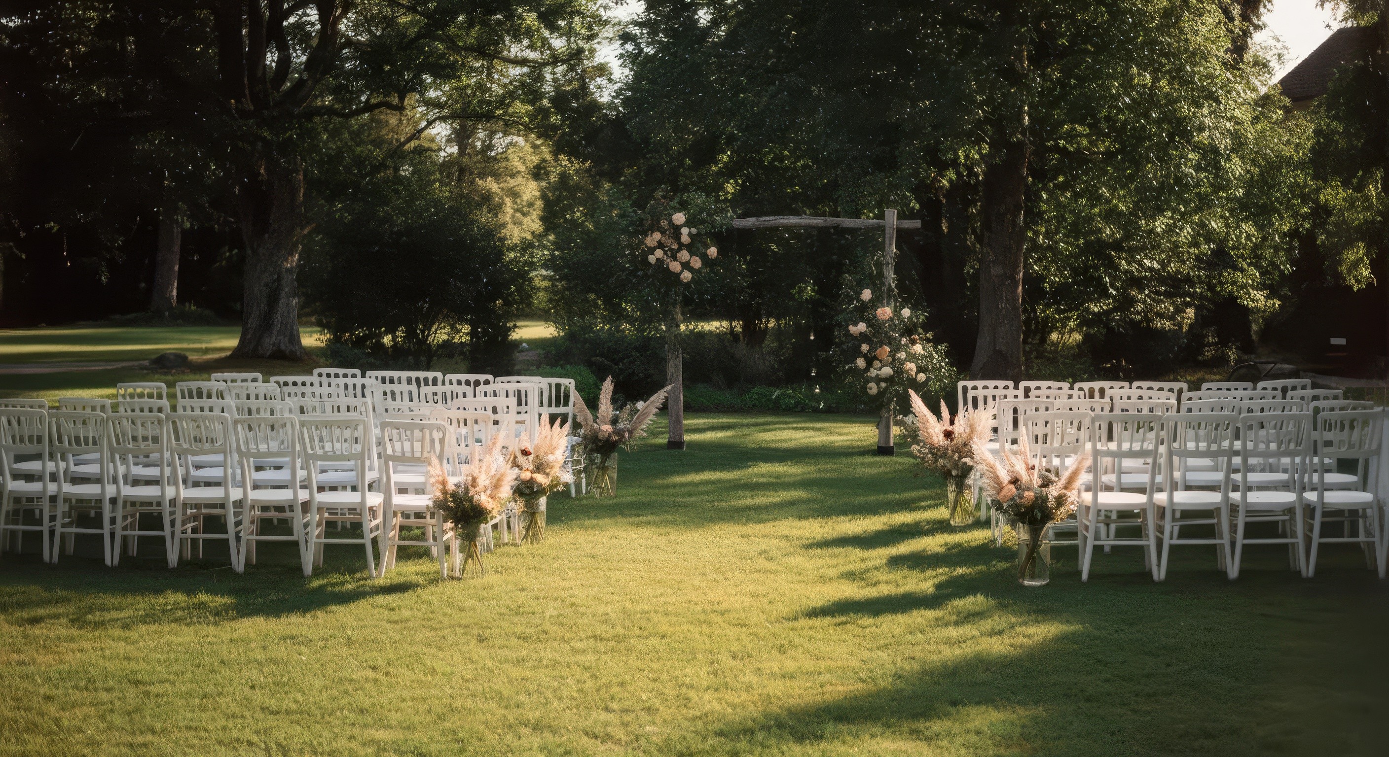 A serene outdoor wedding setup with white chairs arranged on green grass under a canopy of trees.