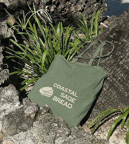 Canvas tote bag laid on the side of a palm tree that says Coastal Sage Bread in sage green with an abstract loaf of bread logo