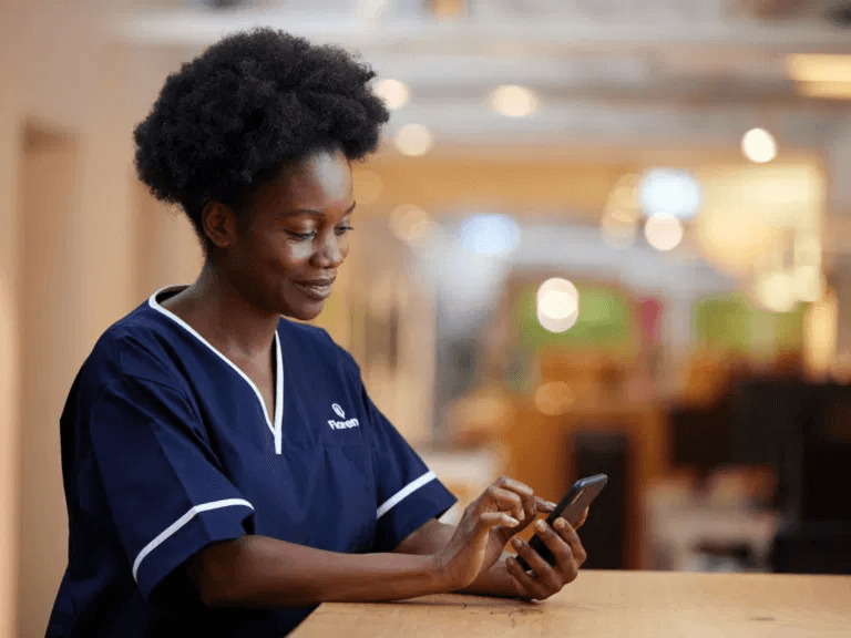 Healthcare worker in scrubs using the Florence app to block bookings while standing at a counter in a warmly lit indoor space.