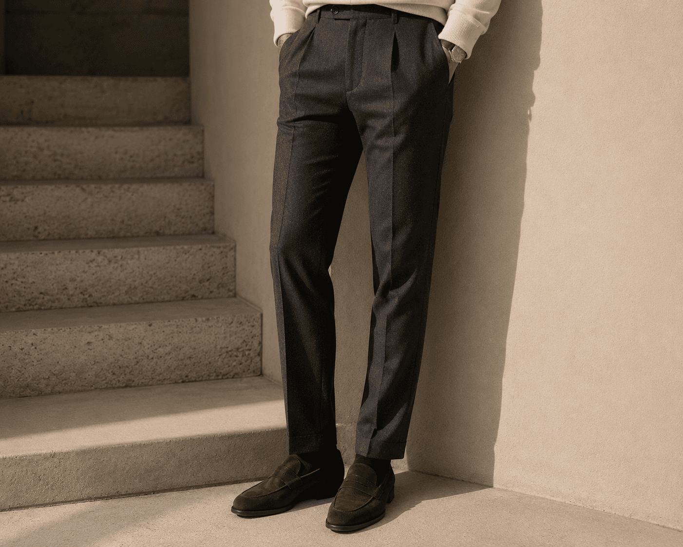 Sided low-angle shot of a man's lower half leaning against a light beige stucco wall next to a set of stone steps. He is wearing high-waisted, pleated charcoal gray flannel trousers with sharp pressed creases, paired with dark brown suede penny loafers and dark dress socks. The bottom hem of an off-white knit sweater and a watch with a leather strap are partially visible.