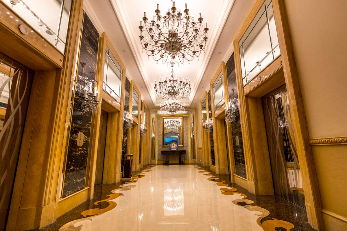 A long hotel hallway with polished marble floors, ornate chandeliers, and gold-trimmed doorways under warm lighting.