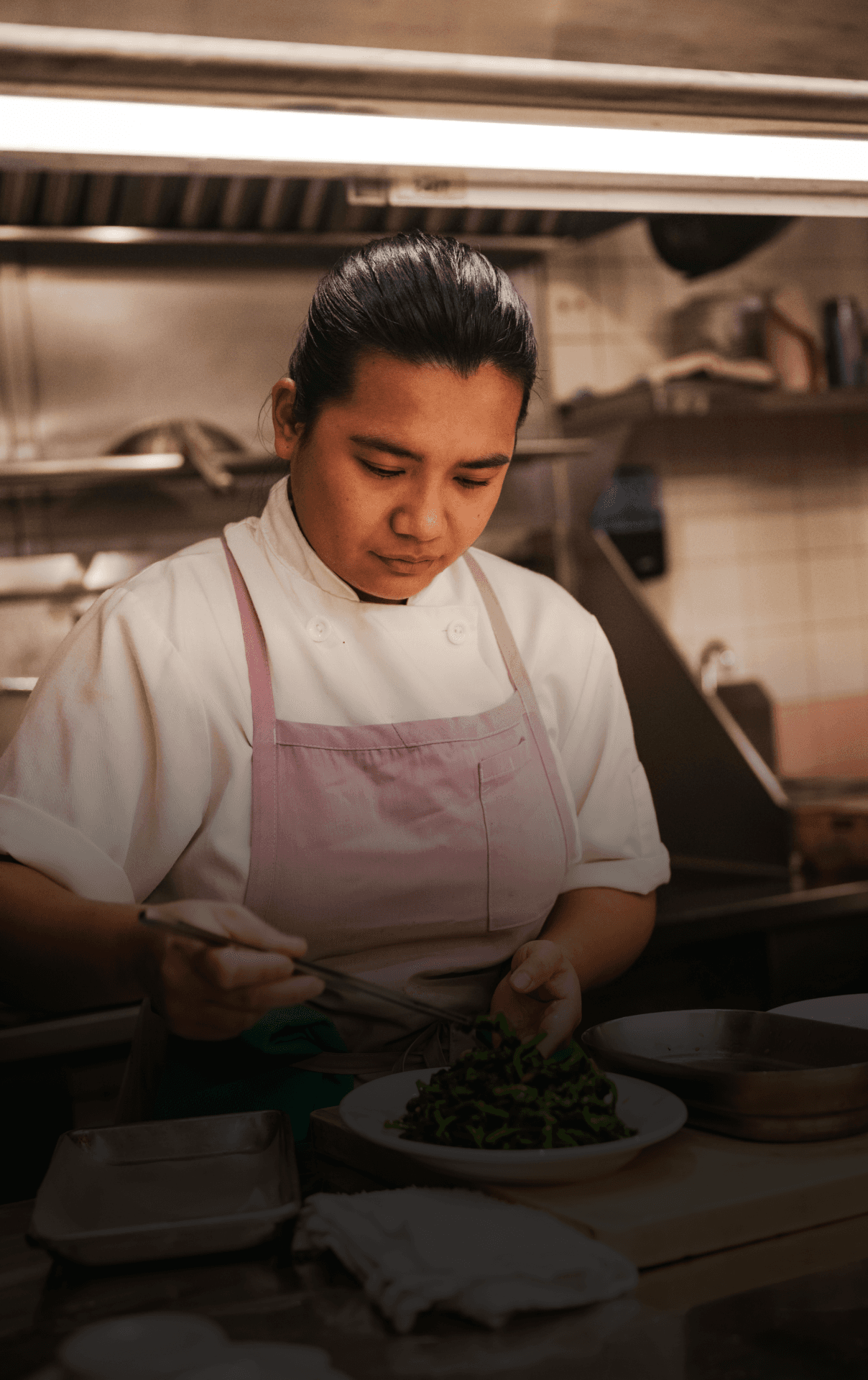 Photograph of a woman wearing an apron at work in a restaurant kitchen, standing over a prep table