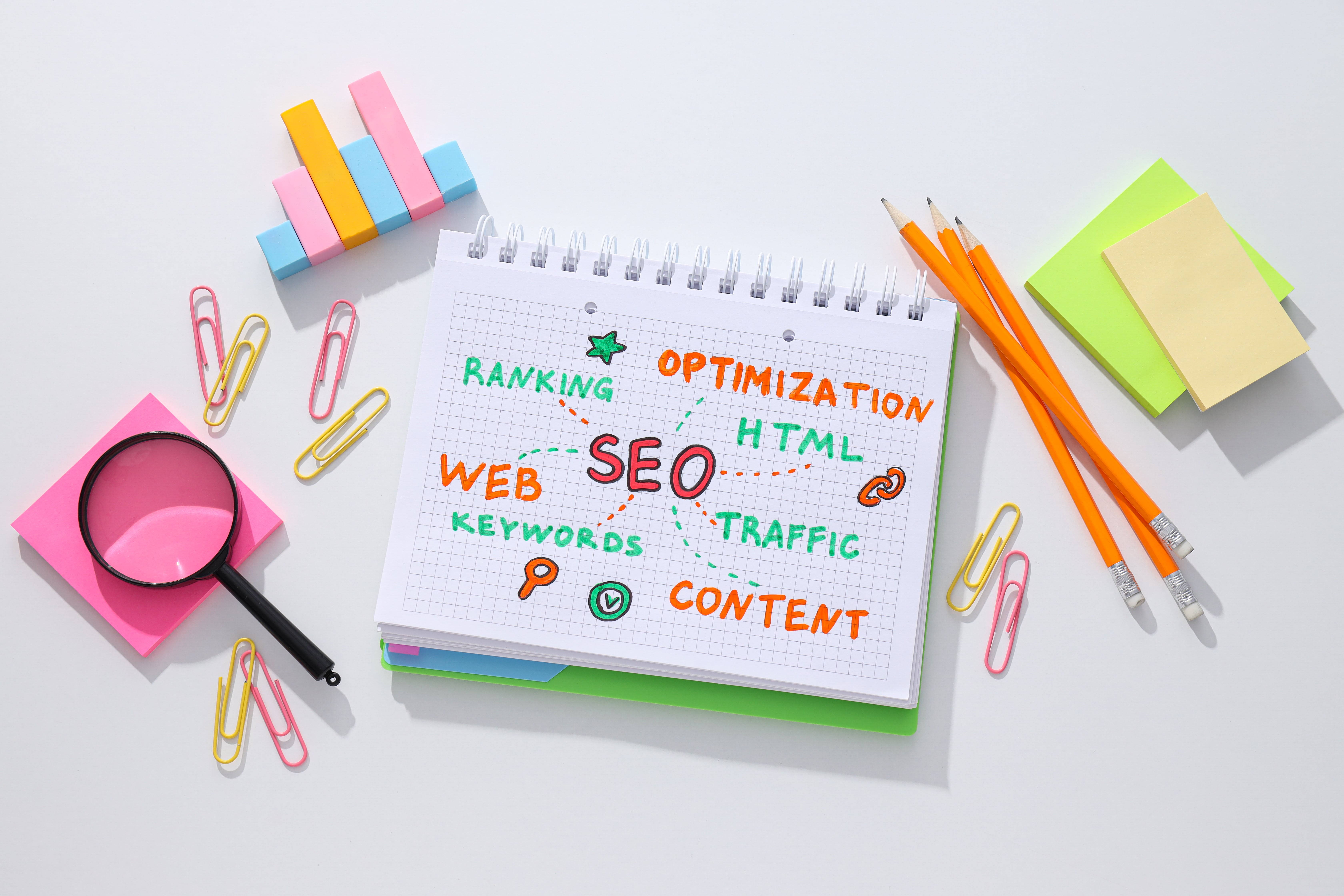 Colorful desk setup with a notebook displaying SEO-related terms like 'Optimization,' 'Traffic,' and 'Content' in bright colors, surrounded by pencils, paperclips, and a magnifying glass, symbolizing the elements of effective search engine optimization.