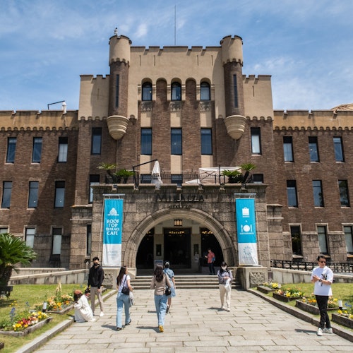 人们走在人行道上，经过一座大型的历史悠久的砖砌建筑，这座建筑有一个中央拱形入口、悬挂的横幅和修剪整齐的草坪。