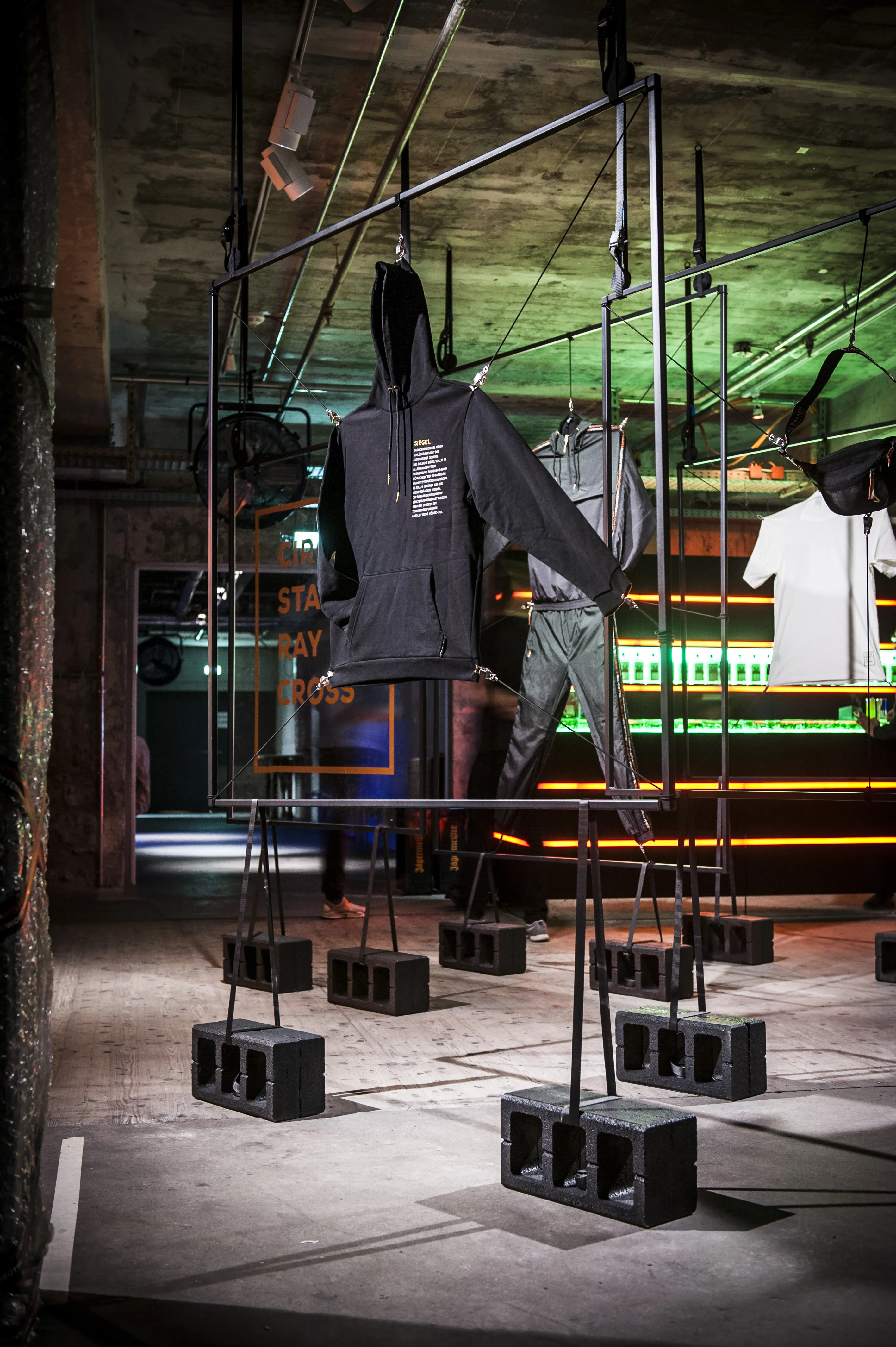 Dark hoodie and tracksuit hanging from a black steel frame weighted with breeze blocks at the JÄGERMEISTER streetwear launch, with an orange-lit bar behind.