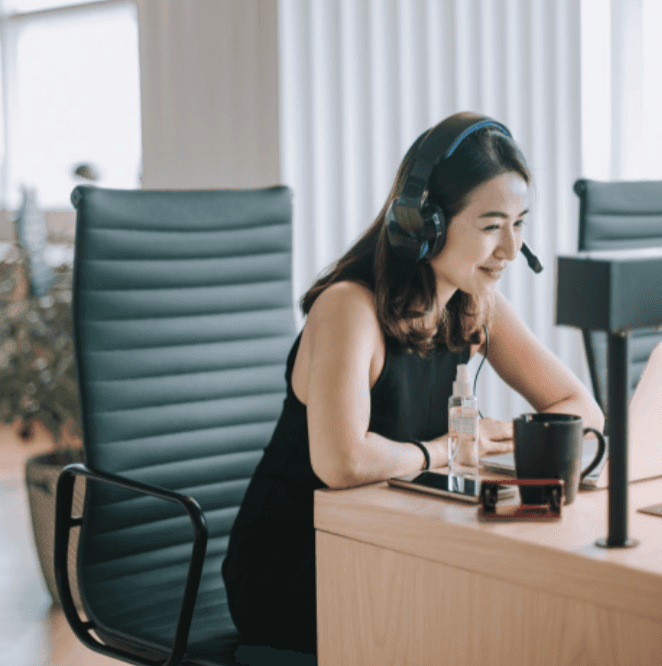 A woman with a headset sits at a desk, looking at her laptop with a coffee cup nearby in a modern office setting.