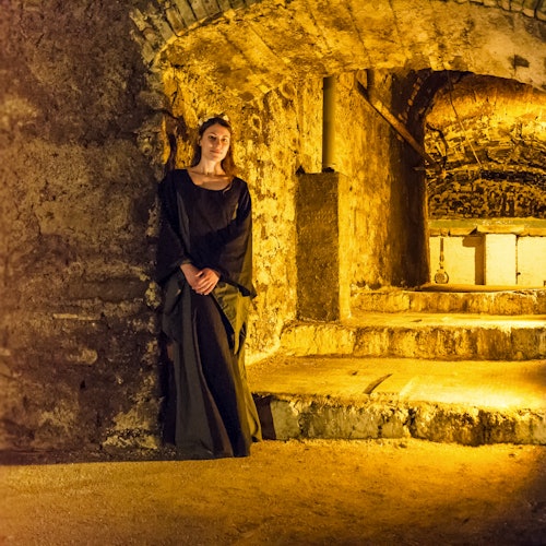 一位身穿深色长裙的女子站在一个昏暗的石墙通道中，通道有拱门和一个被温暖灯光照亮的楼梯。