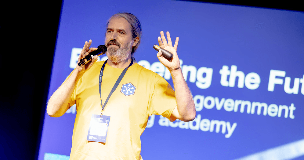 A speaker in a yellow conference t-shirt stands on stage holding a microphone and presentation clicker while delivering a talk. A large blue presentation screen is visible in the background with partially visible text about a government engineering academy. The speaker is wearing a conference lanyard and is gesturing with both hands during the presentation.