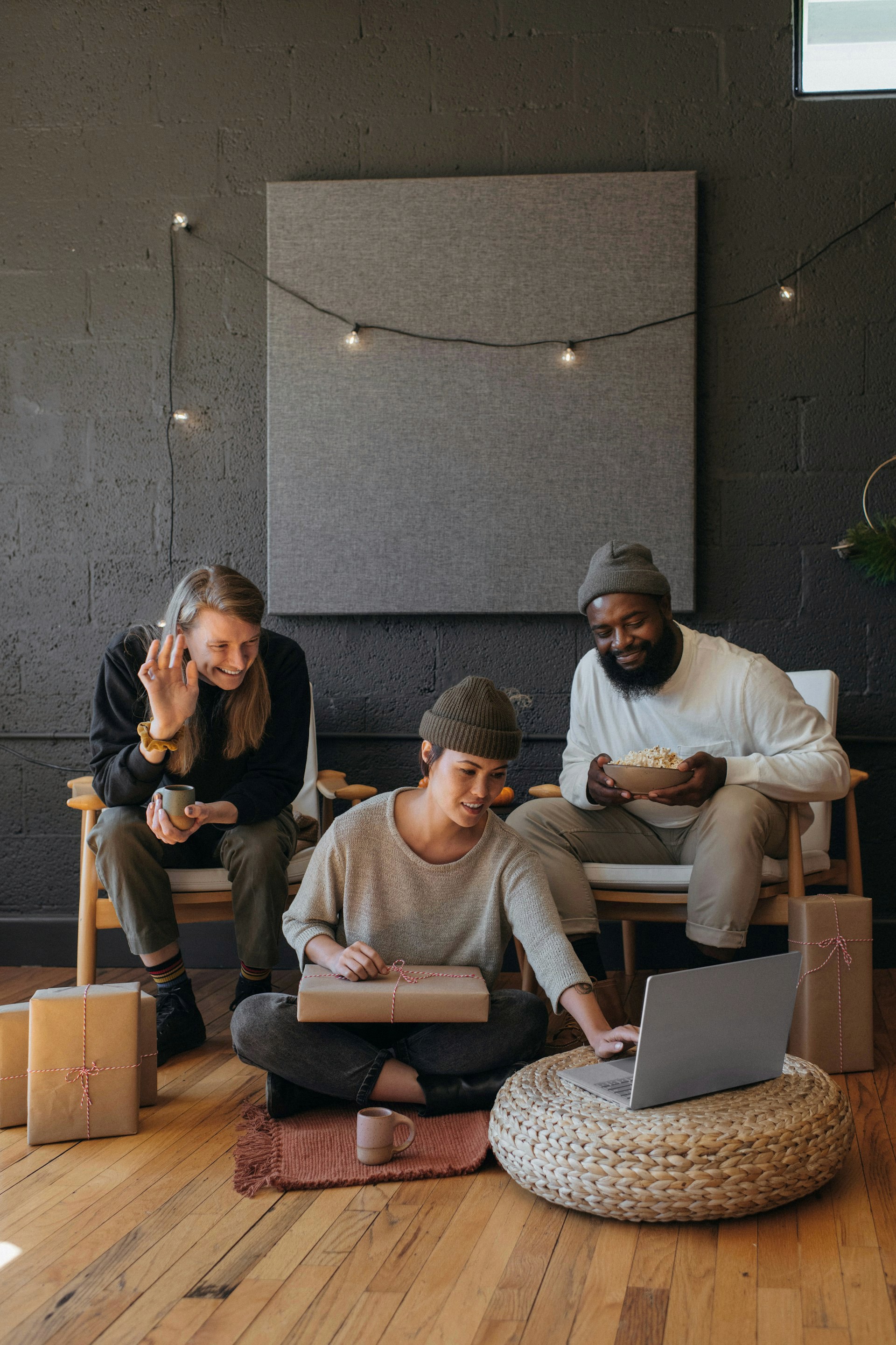 Three friends celebrating with gifts and a laptop; one sits on a pouf, the others on chairs.