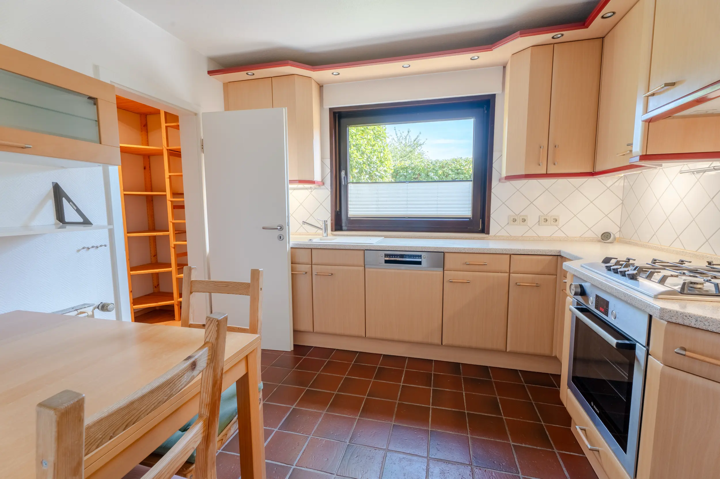 Einbauküche mit hellen Holzfronten, Gasherd, Kühlschrank, Fenster und kleinem Essplatz.