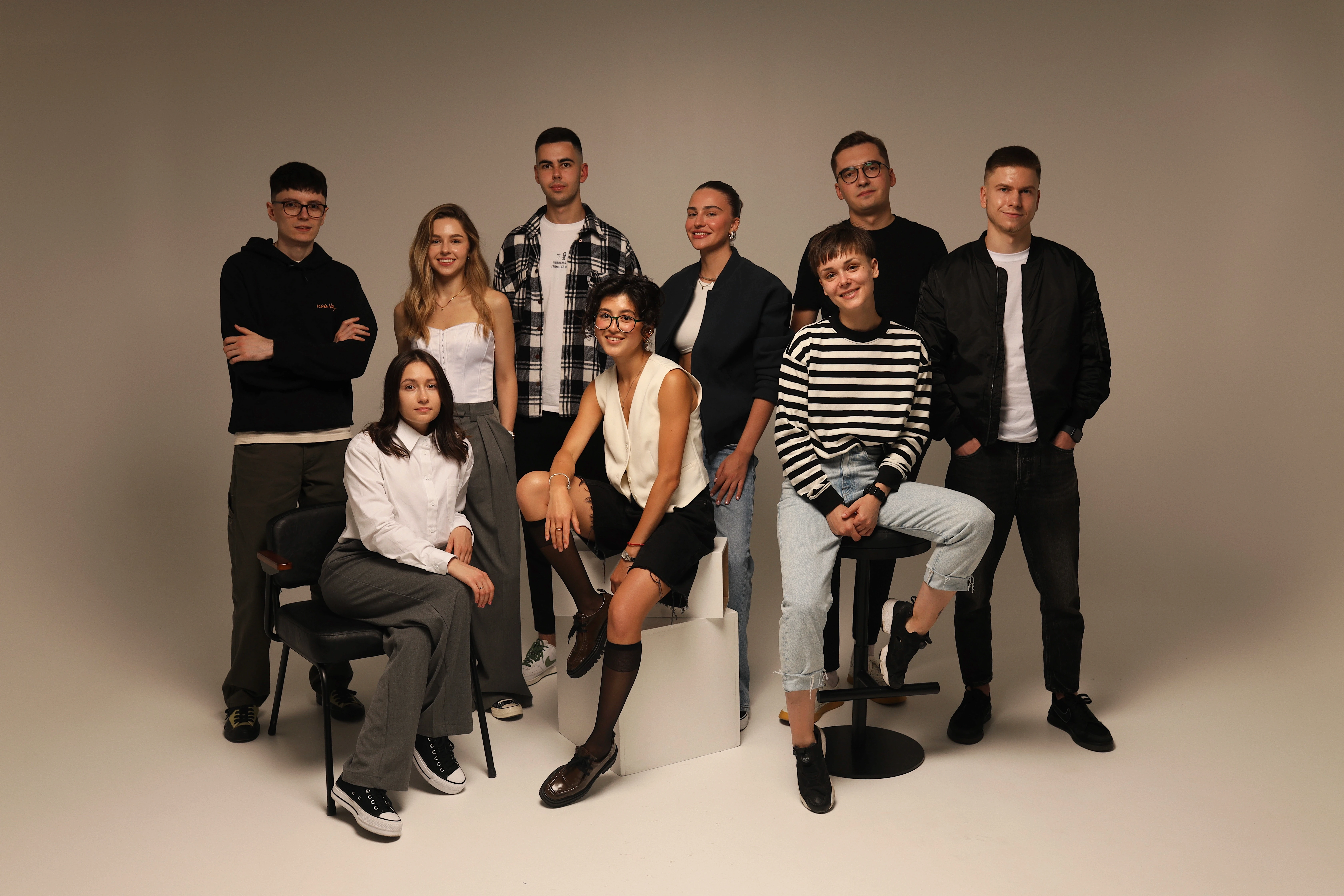 This image shows a group of eight young adults posing for a photo in a studio with a neutral background. They are stylishly dressed in a mix of casual and smart-casual clothing. Four women and four men are arranged in a relaxed, yet organized fashion. Two are seated on blocks, one is seated on a chair, and the rest are standing. Their expressions are pleasant and friendly, suggesting a sense of camaraderie or teamwork. The lighting is soft, and the overall mood of the photo is professional but approachable.