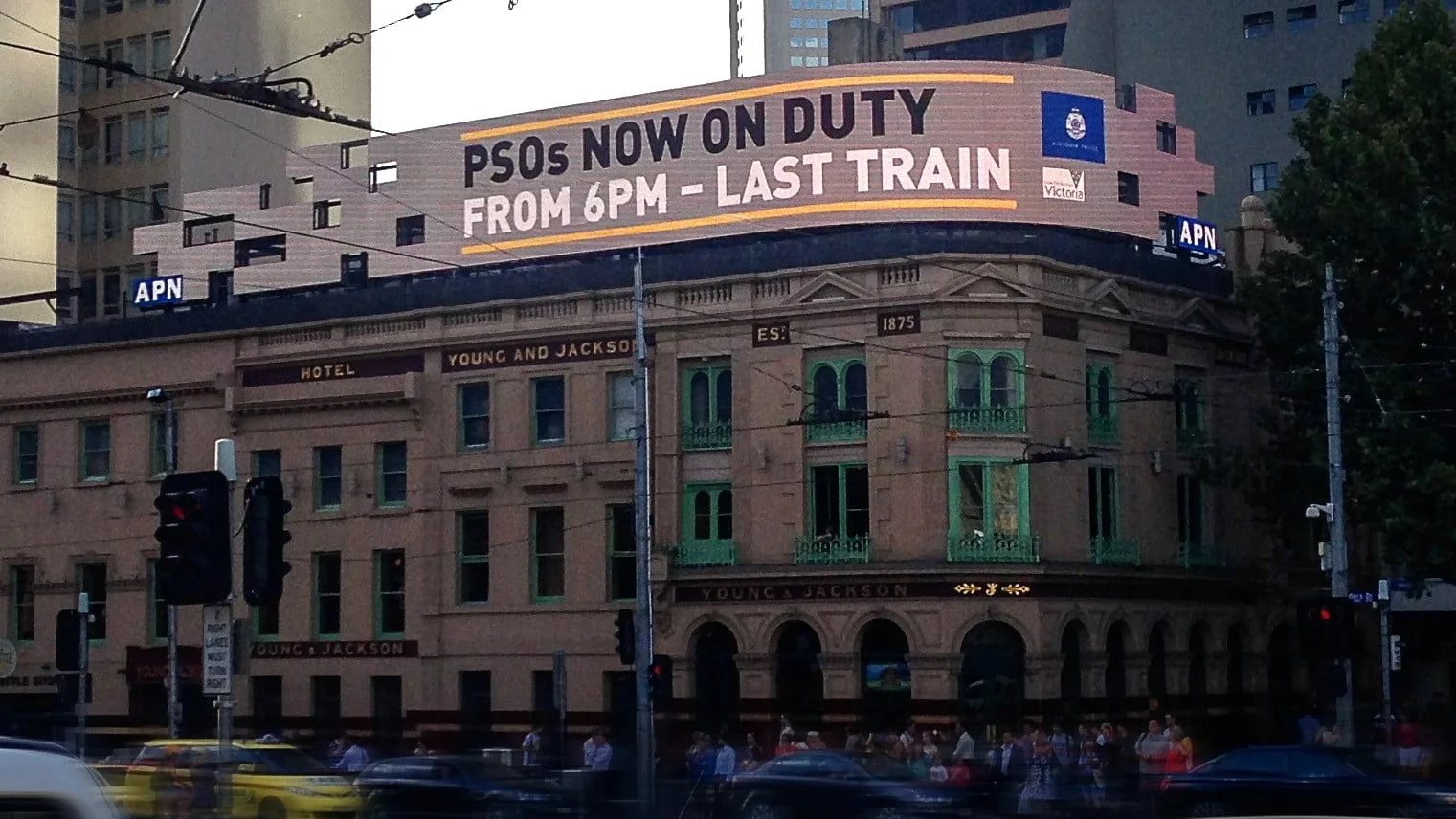 Building rooftop sign reading 'PSOs now on duty from 6pm - last train' for Victoria Police, above a Melbourne pub
