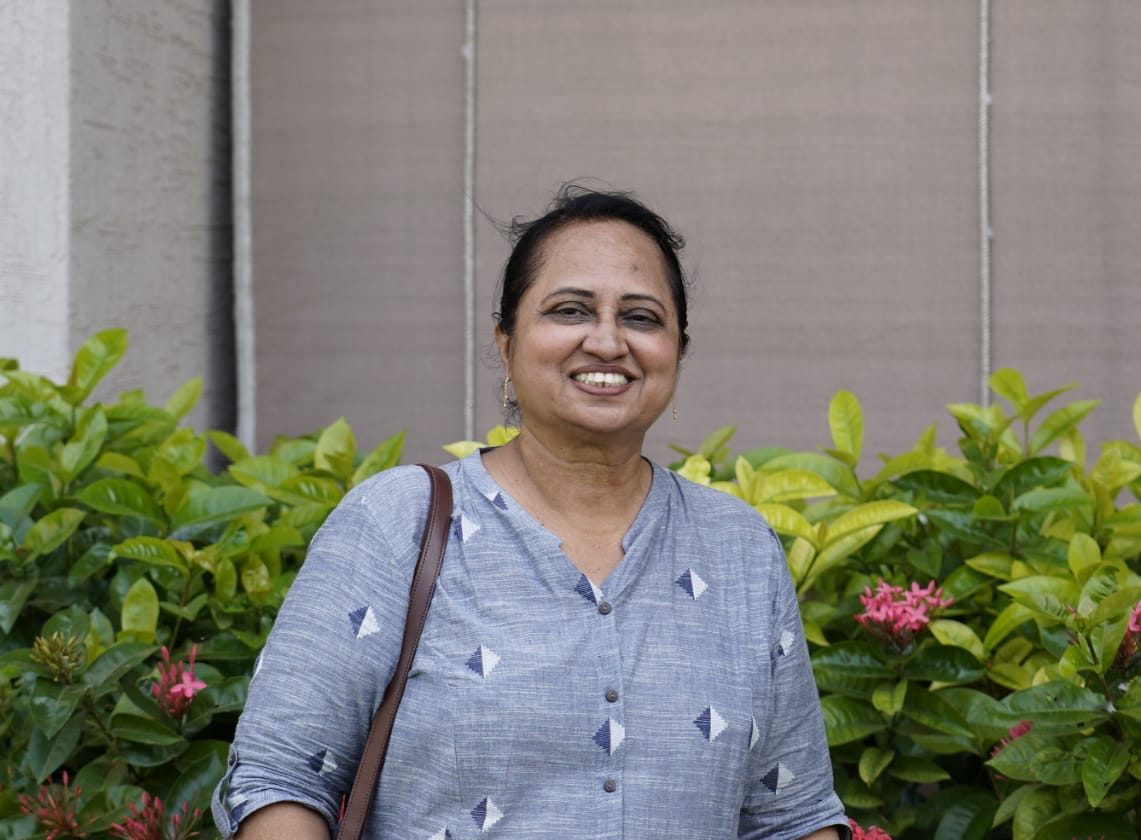 an Indian woman with dark hair a blue shirt and a shoulder bag smiling at camera