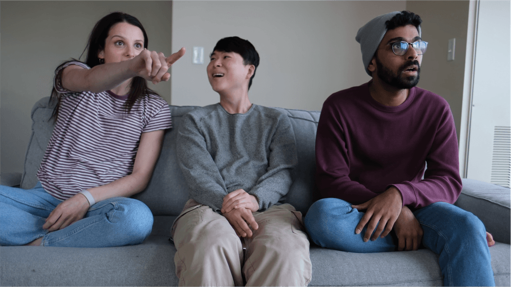 Three people sit on the couch watching TV, pointing at the screen and looking at each other excitedly.