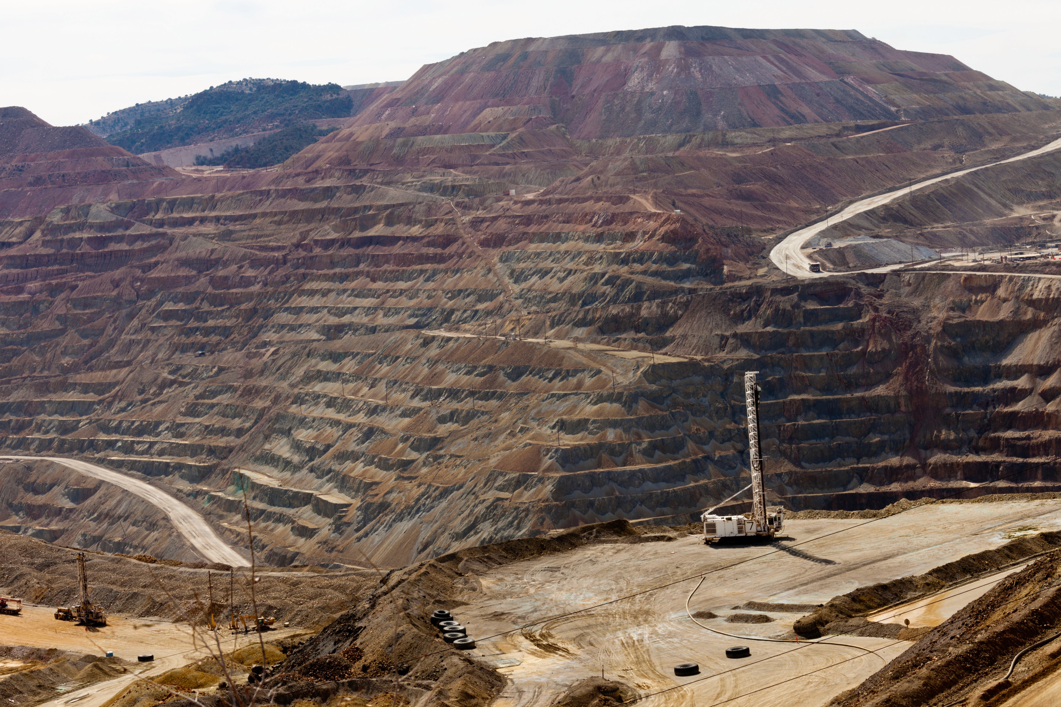 Aerial view of a stepped open-pit mine surrounded by forested hills, with visible machinery and terraced mining levels.