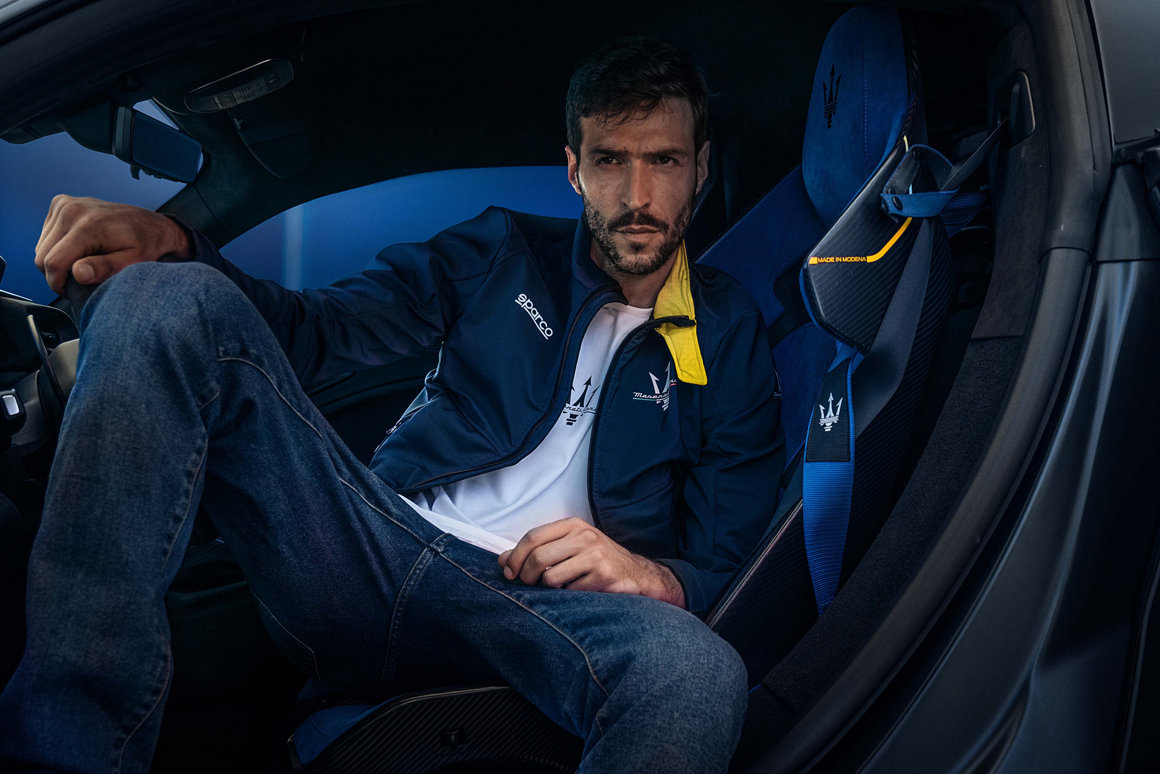 Un homme assis dans une voiture de sport haut de gamme, portant une veste de sport Maserati avec des détails jaunes.