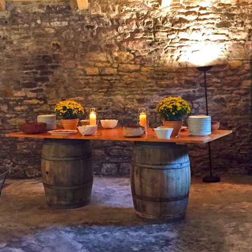 一張由木桶製成的質樸桌子上擺著花、蠟燭、盤子和碗，背景是柔和燈光照射下的紋理磚牆。