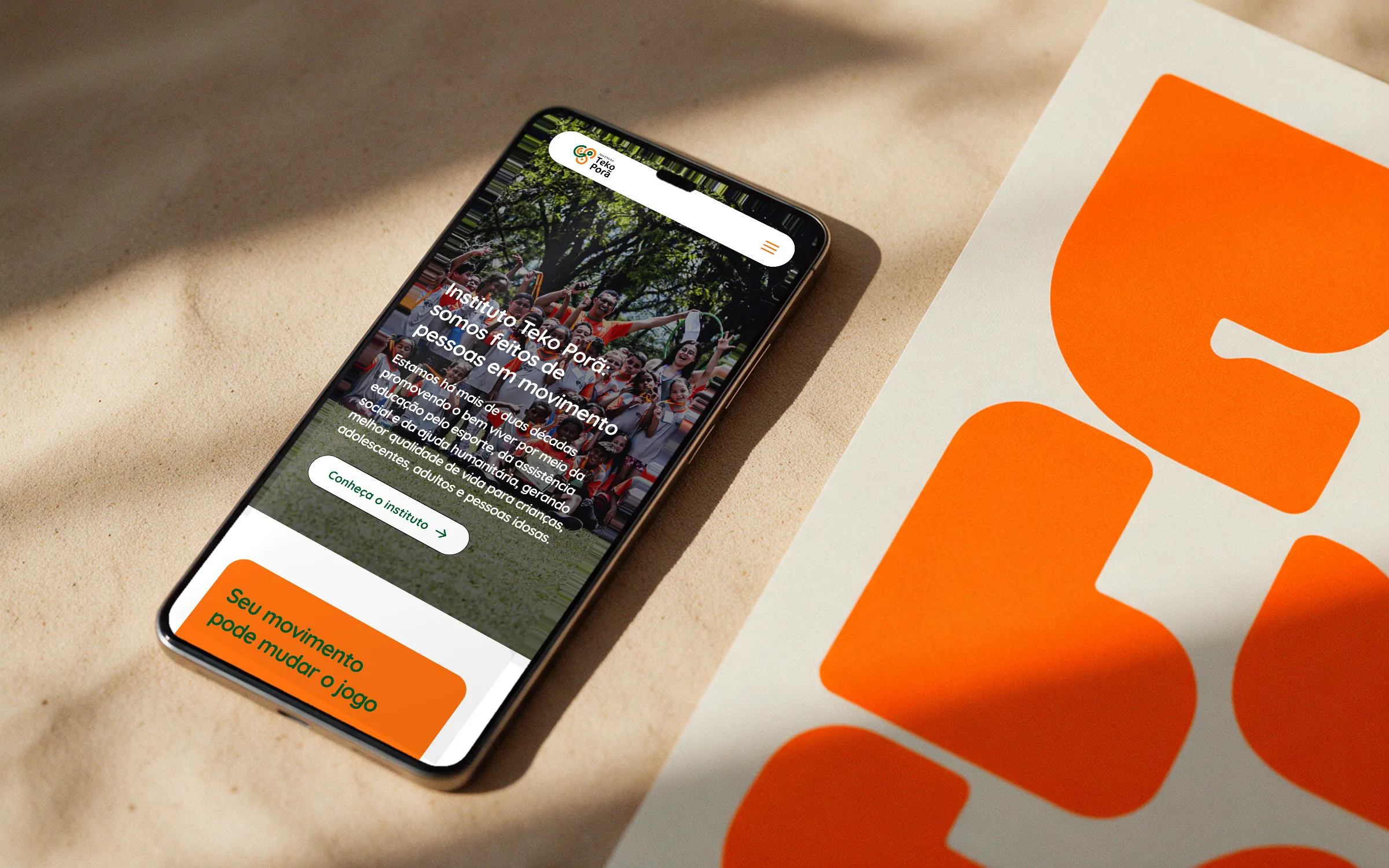 Página inicial do site do Instituto Teko Porã num celular ao lado de um impresso laranja