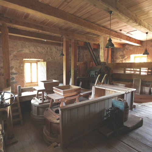 An old mill interior with wooden beams, various milling equipment, and sunlight streaming through a window.