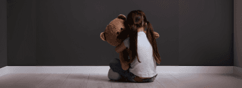 Child sitting alone hugging a teddy bear in a dim room.