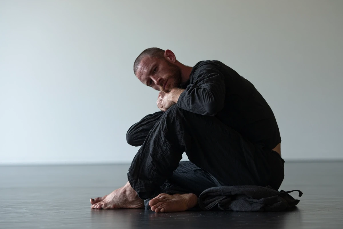 A person sitting against a wall with one hand tucked under the opposite armpit in a self-hug position, both feet pressed firmly against the floor.