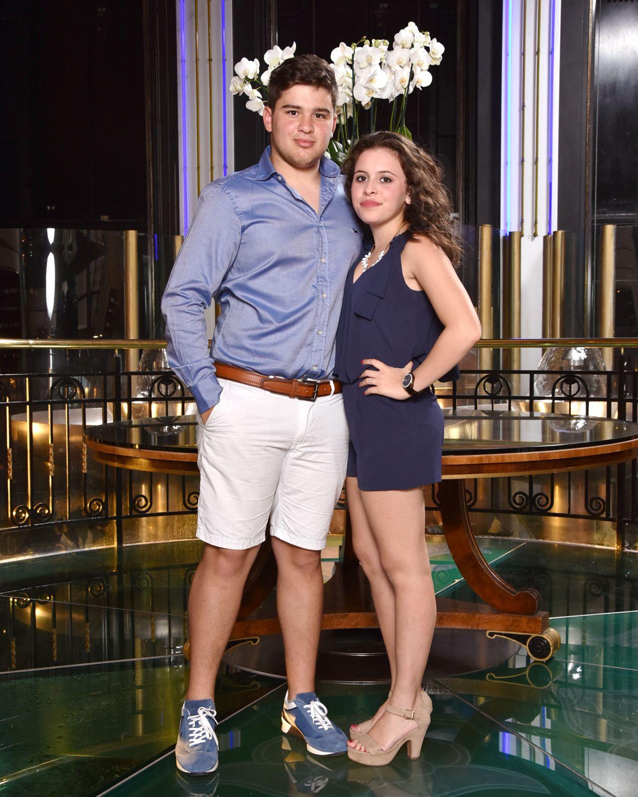 A couple stands on a sandy hill at sunset, facing away, with arms raised in a relaxed "hang loose" gesture. The scene conveys tranquility and joy.