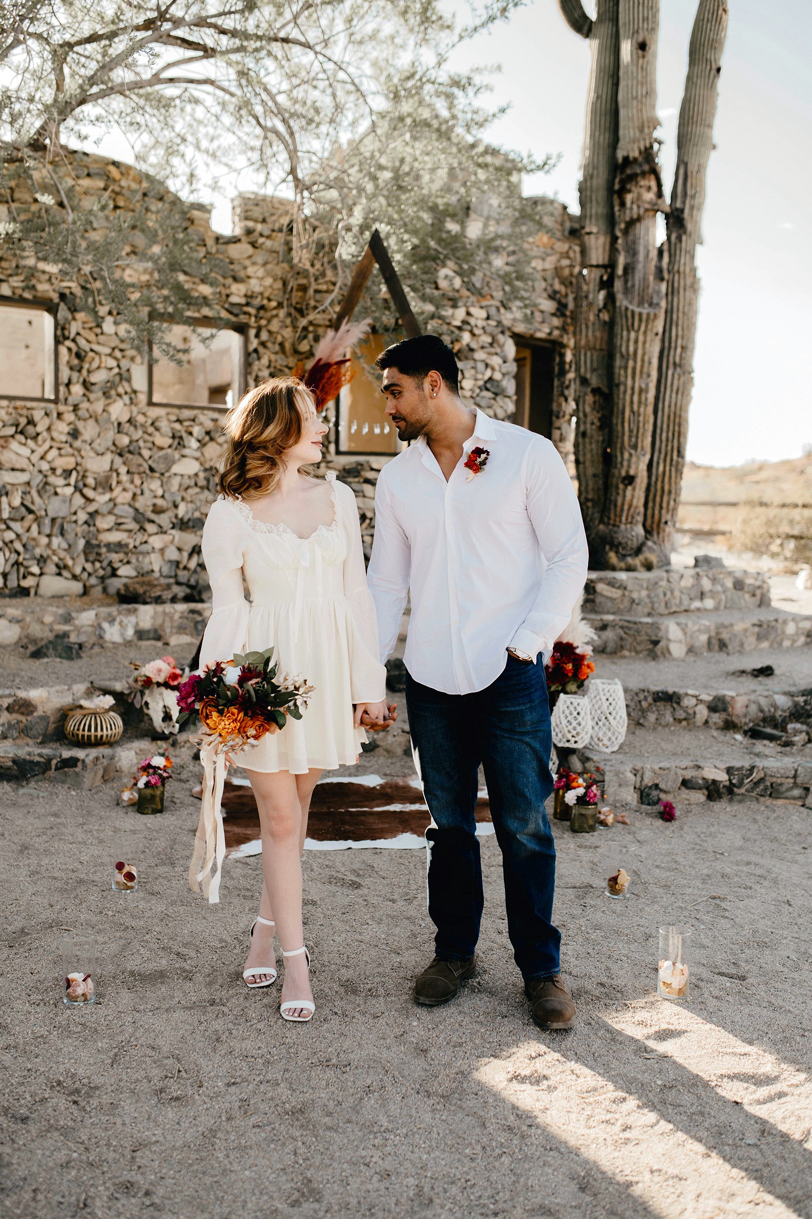 A couple stands hand-in-hand in front of a rustic stone building.