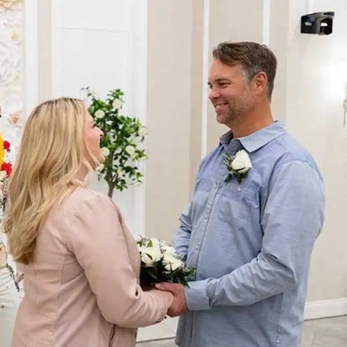 En man och kvinna ler mot varandra, håller varandras händer, båda bär blomsterknappar i en dekorerad inomhusmiljö.