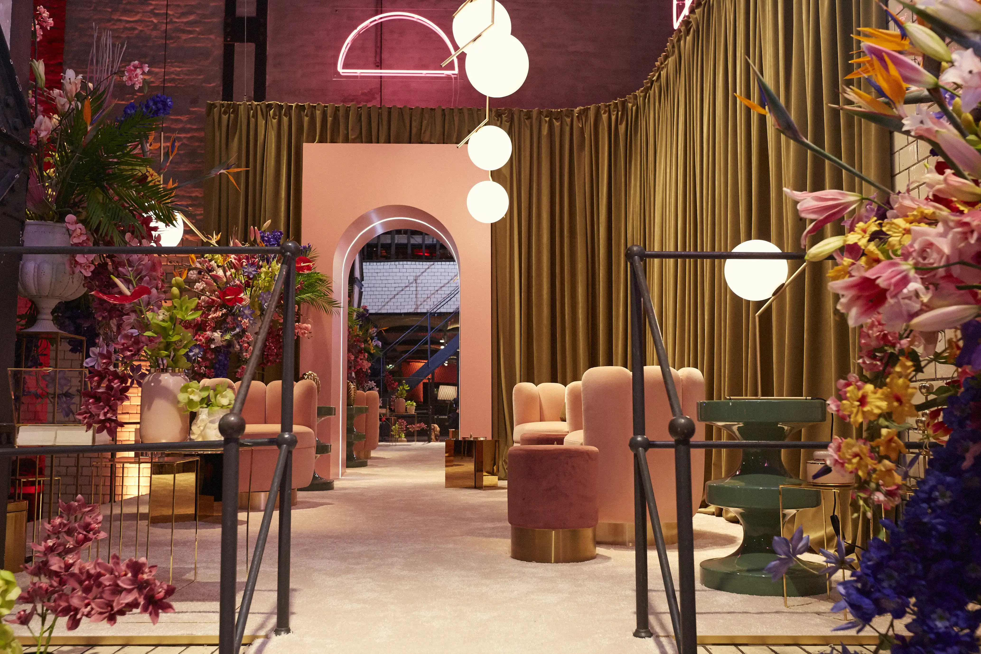 Pink lounge of the Klarna Fashion Week pop-up with velvet armchairs, an arched passage, olive curtains and abundant flower decor.
