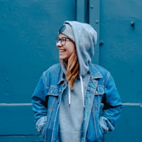 A smiling person wearing glasses, a gray hoodie, and a blue denim jacket stands with hands in pockets against a blue wall.