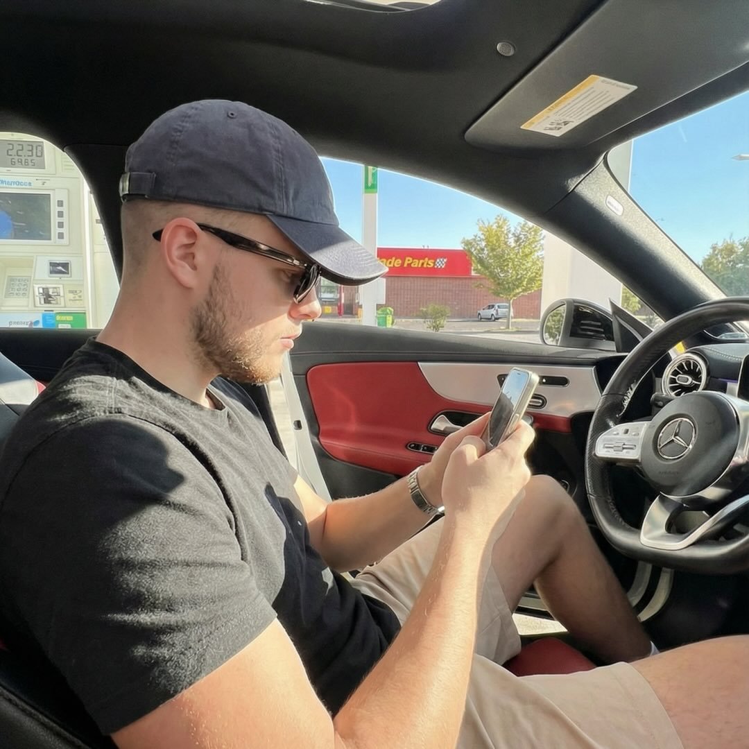 A young man with light brown hair is wearing a black suit jacket and white shirt, standing in a softly lit office setting, conveying professionalism.