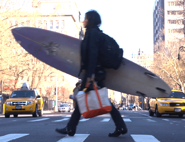 A person with dark hair walks across a crosswalk in a city, carrying a large, light-colored surfboard under their arm and a white and orange tote bag in their hand. They are wearing a dark jacket and dark pants. The crosswalk is marked with white stripes on the asphalt. Behind them, a yellow taxi is visible, and tall buildings line the street. Bare trees line the sidewalk in the background. The sky is bright and clear.