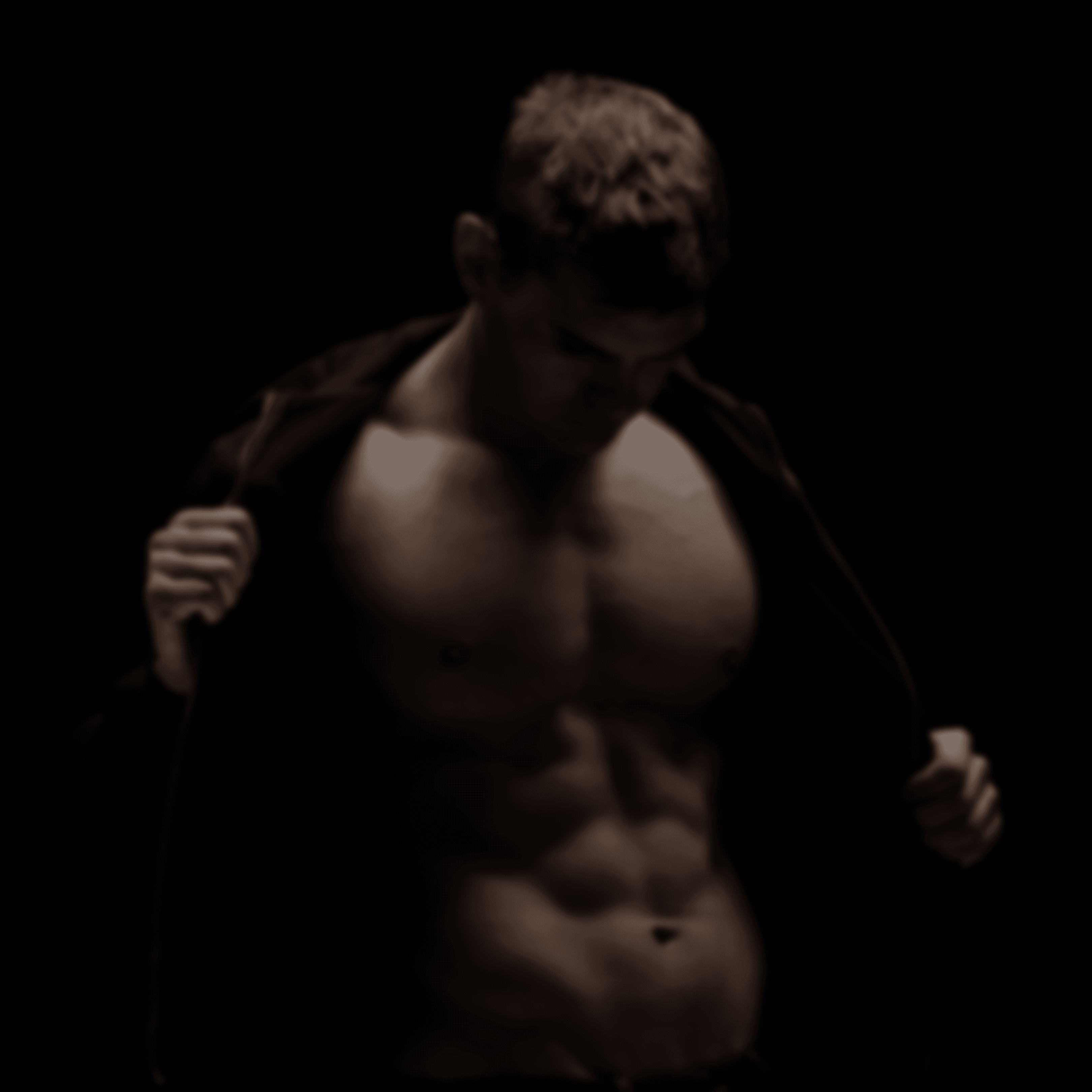 A man posing against a dark backround revealing a naked torso as he unzips his hoodie.
