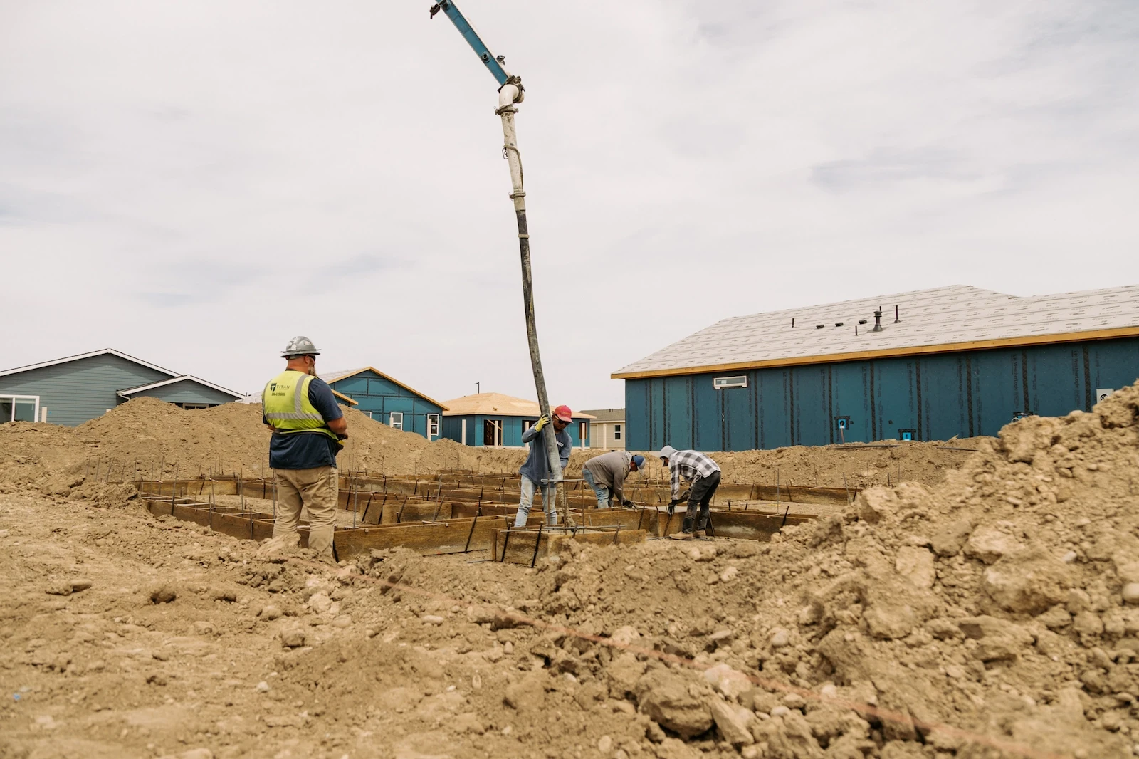 A crew of workers guides a concrete pump hose to pour footings into rebar-reinforced forms, while a supervisor in a safety vest looks on at a residential construction site.