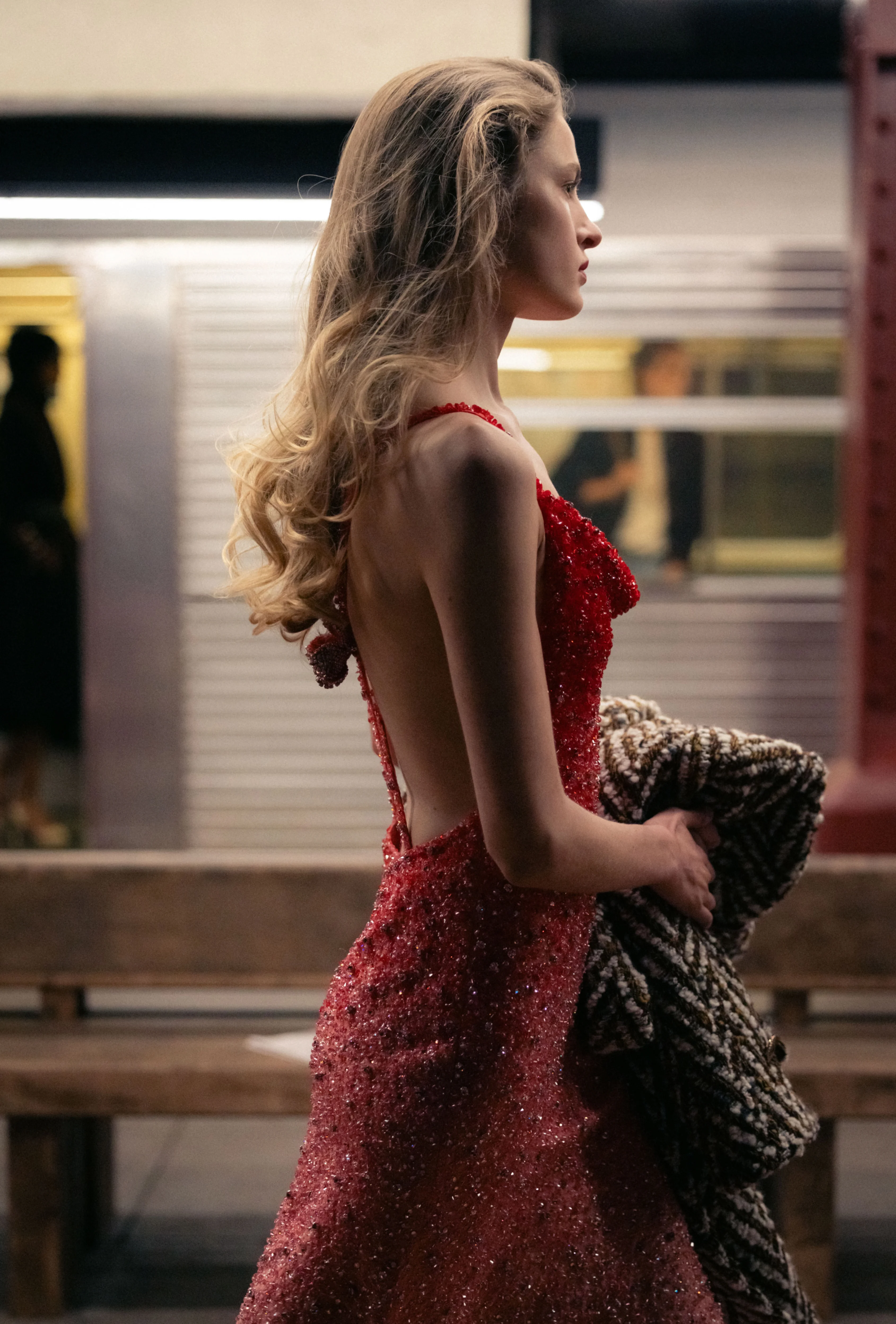 A model in a red sequined gown carries a textured coat while standing on a subway platform at the Chanel Métiers d’Art 2026 show.