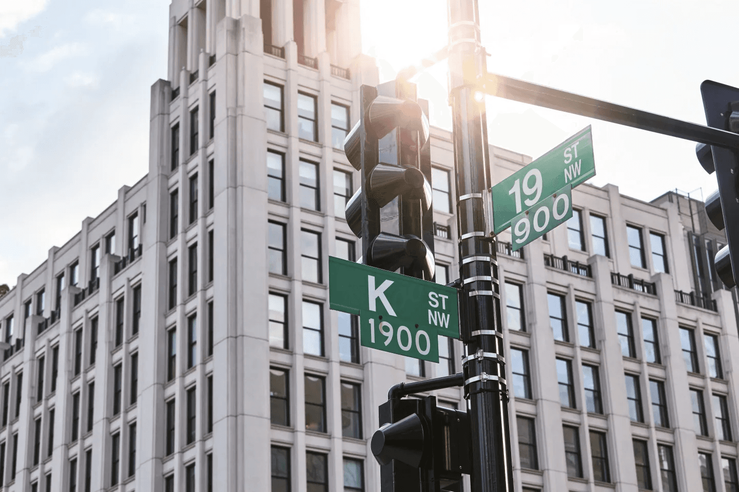 Street signs on a traffic signal in front of a tall building, with sunlight shining in the background.