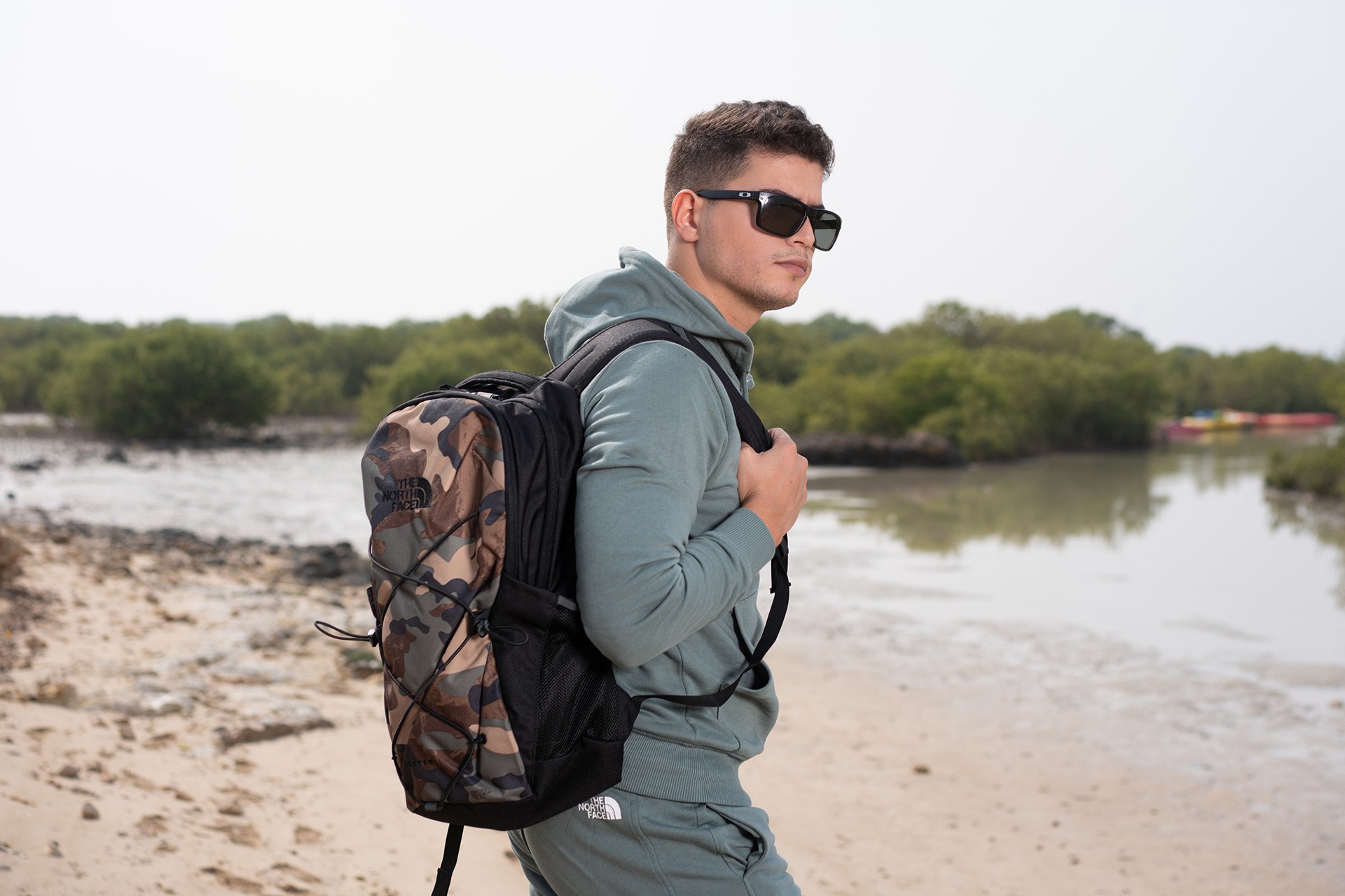 A man in a teal hoodie and sunglasses with a backpack, walking on a sandy shoreline edged with mangroves.