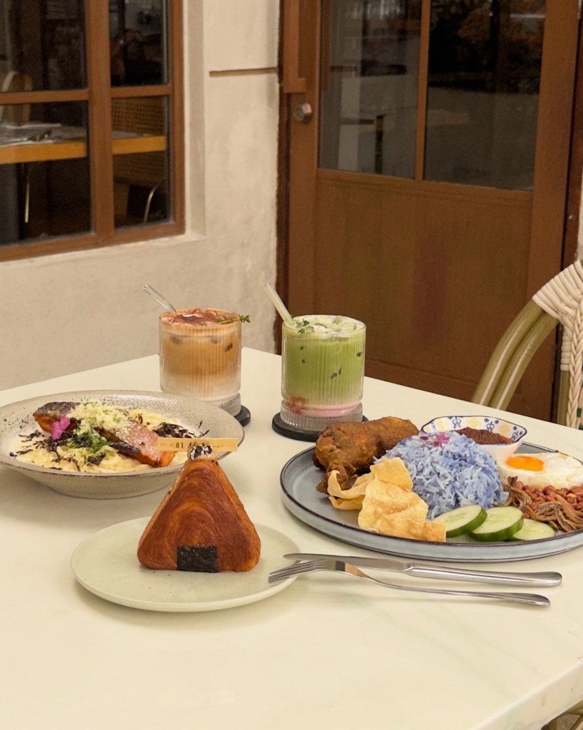 A table setting featuring various dishes, including a salmon plate with creamy sauce and greens, an onigiri-shaped croissant, a plate of fried chicken with blue rice, cucumber slices, and crackers, alongside two drinks: a brown iced beverage and a green one with cream.