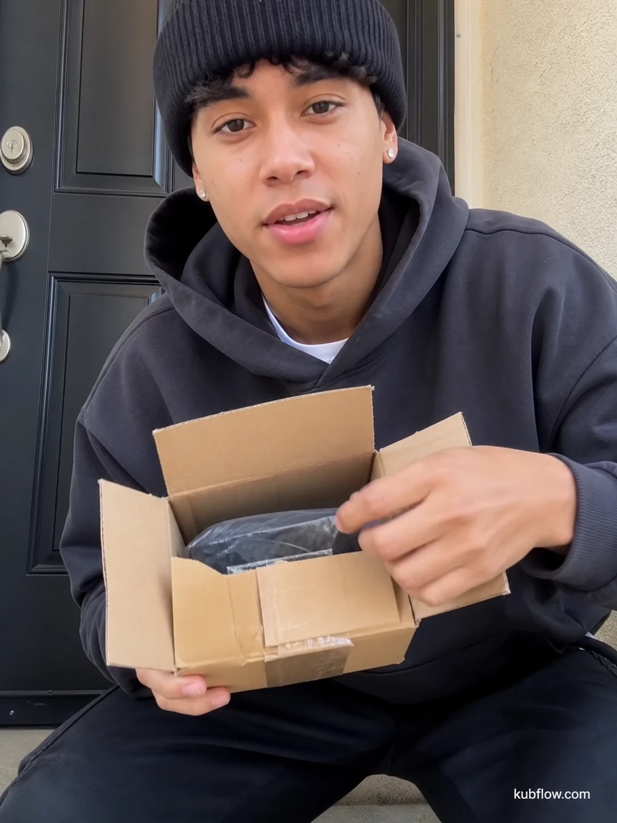 UGC-style photo: streetwear creator, by a front door before going out, unboxing a small generic package while talking to camera, casual handheld angle.